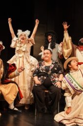 Photograph of the six performers of Monarchs Anonymous on stage, in costume. The monarchs pose enthusiastically around Dr Thompson, who is sitting down.